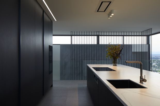 A sophisticated modern kitchen with dark full-height cabinetry, a large pale stone island with integrated cooktop and sink, and a sculptural curved tap. A vertical metal screen separates the kitchen from the glazed exterior, while linear ceiling lighting and concealed appliances reinforce the minimalist design. A vase of foliage provides a small natural accent, and a distant city view is visible through the opening.