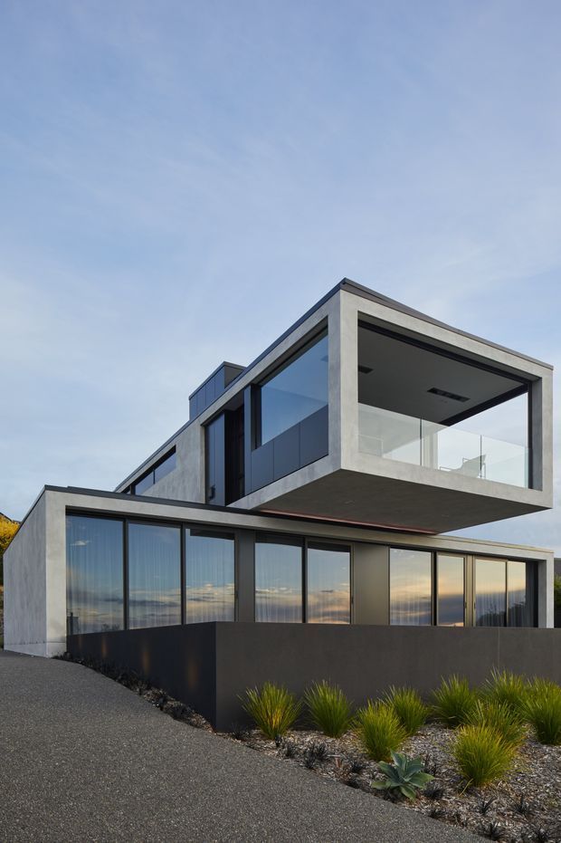 Exterior view of a contemporary multi-level residence with strong geometric forms, exposed concrete framing, extensive dark-framed glazing, and a cantilevered upper-floor balcony. The home is set above a landscaped frontage with gravel, grasses, and low planting.