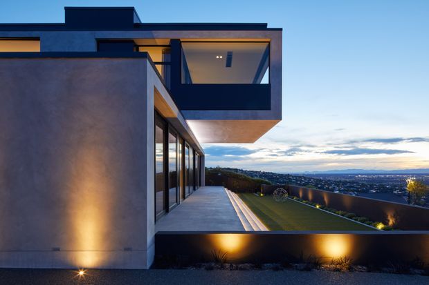 Contemporary multi-level residential exterior at dusk, featuring smooth rendered walls, dark flat roof forms, extensive floor-to-ceiling glazing, and a projecting upper balcony or cantilevered volume. Warm architectural lighting illuminates the facade, terrace, retaining wall, and landscaped garden, with an expansive suburban or urban outlook beyond.