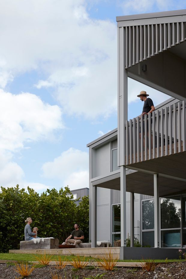 Exterior architectural photograph of a contemporary multi-level home with pale grey cladding, strong rectangular structural framing, and projecting balconies enclosed by vertical slatted screens. A person stands on the upper balcony, while two people sit in an outdoor lounge area beside low landscaped planting. Large glazed doors and windows open toward the garden, with a bright partly cloudy sky behind the building.