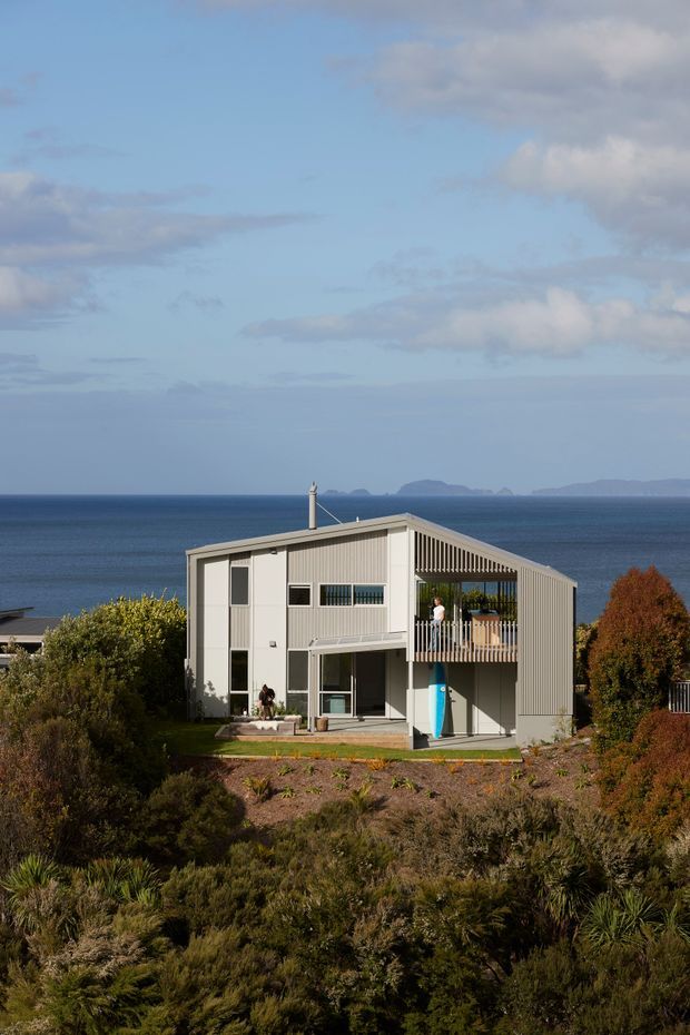 A contemporary two-storey coastal house set within native landscaped vegetation, photographed from the front. The home has a simple rectangular form with light grey corrugated metal cladding, a steeply angled roof or upper facade, large glazed openings, a sheltered upper balcony with vertical screening, and a surfboard stored beneath it. The ocean and distant islands are visible behind the property.