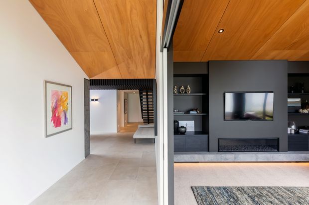 A contemporary open-plan interior viewed through a large central glazed opening, showing a hallway and living room. The living room features a dark built-in media wall with television, fireplace and shelving, while the hallway has pale tiled flooring, white walls, and a timber-lined ceiling extending across both spaces. A staircase and framed artwork are visible in the background.