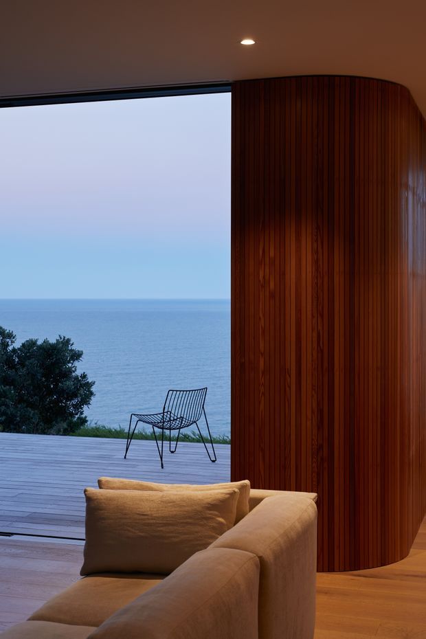 A refined coastal living room opens directly onto a timber deck and expansive ocean view through floor-to-ceiling glazing. The interior features a large curved caramel upholstered sofa, warm timber flooring, and a dramatic curved wall clad in vertically ribbed dark timber panelling. A minimalist outdoor chair and coastal vegetation are visible beyond the opening, creating a warm, serene indoor-outdoor composition.