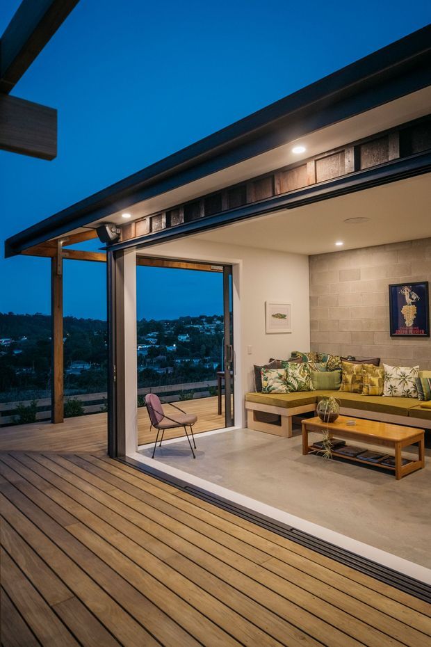 Evening view of a contemporary indoor-outdoor living area opening onto a broad timber deck. Large sliding glass doors connect the deck to a living room with a built-in sectional sofa, patterned cushions, timber coffee table, exposed blockwork feature wall, and recessed ceiling lighting. The home overlooks a suburban hillside landscape.