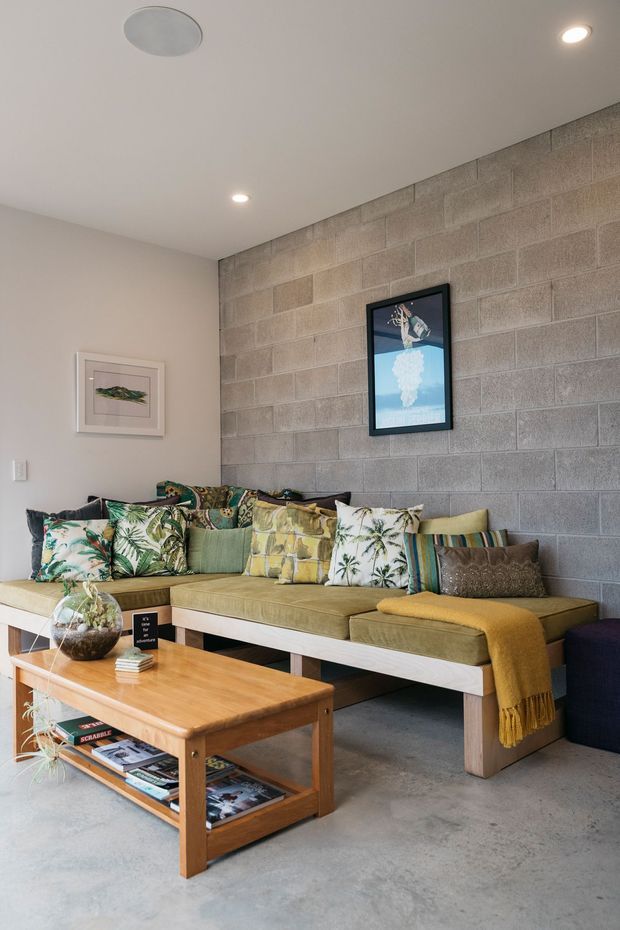 A compact, inviting living room with a built-in sectional sofa against an exposed grey concrete block feature wall. The room has a smooth white ceiling with recessed downlights, a polished-looking concrete floor, a timber coffee table, tropical botanical cushions, and a mustard throw. Framed artwork and a few indoor plants add decorative detail.