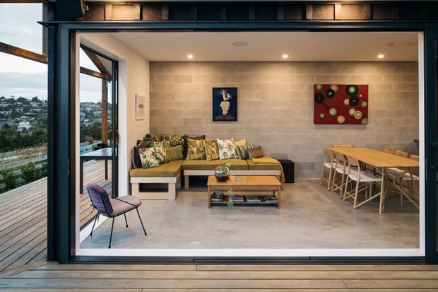 Contemporary indoor-outdoor living and dining area opening onto a timber deck through expansive dark-framed sliding glass doors. The room features a pale rendered masonry feature wall, polished concrete floor, built-in sectional seating with patterned cushions, timber coffee table, and a timber dining table with woven chairs. A green hillside suburb is visible beyond the deck.