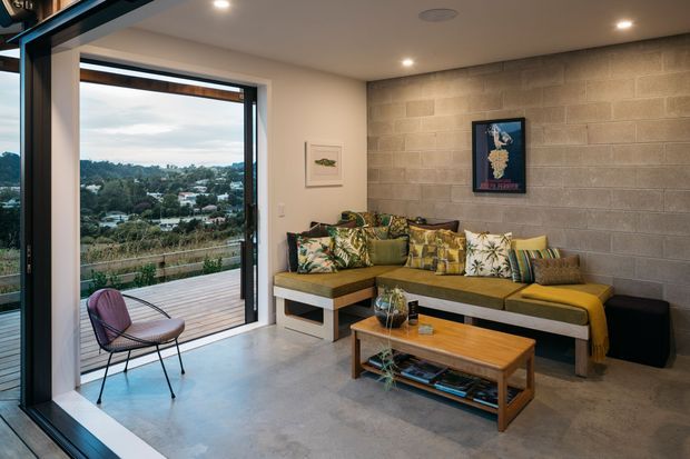 A contemporary living room with polished concrete flooring, a grey concrete-block feature wall, and a built-in L-shaped sofa layered with colourful botanical cushions. Large sliding glass doors open onto a timber deck and provide expansive views over a leafy suburban landscape. A timber coffee table, artwork, recessed ceiling lights, and a sculptural lounge chair complete the space.