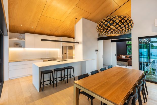 Open-plan contemporary kitchen and dining area with white built-in cabinetry, a large island with bar stools, timber flooring, and a warm timber-lined ceiling. A long solid timber dining table sits beneath a prominent woven black pendant light, with glazing and a living area visible beyond.