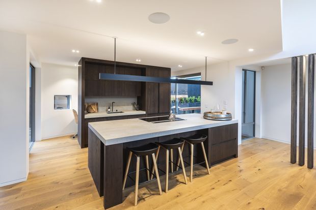 A contemporary open-plan kitchen with a large dark timber island, pale stone benchtop and seating for four. The kitchen includes dark timber cabinetry, integrated appliances, a linear black pendant light, white walls, recessed ceiling lights and warm natural timber flooring. Glazing and a doorway provide views toward an outdoor area, while vertical dark timber battens define the right side of the space.