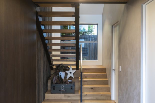 Interior view of a compact modern stairway and hallway. Open-riser timber stairs ascend beside a dark timber-clad wall, with black metal balustrade and handrail. A lower flight leads toward a bright landing with a large window, while a white door and pale textured walls line the corridor. Warm timber tones contrast with dark metal and neutral plaster finishes.