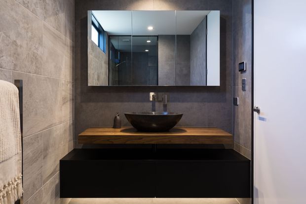 Contemporary bathroom vanity featuring large-format grey stone-look wall tiles, a wall-mounted matte black cabinet, a timber benchtop, and a black vessel basin beneath a mirrored cabinet. A white door and towel rail are visible at the sides.