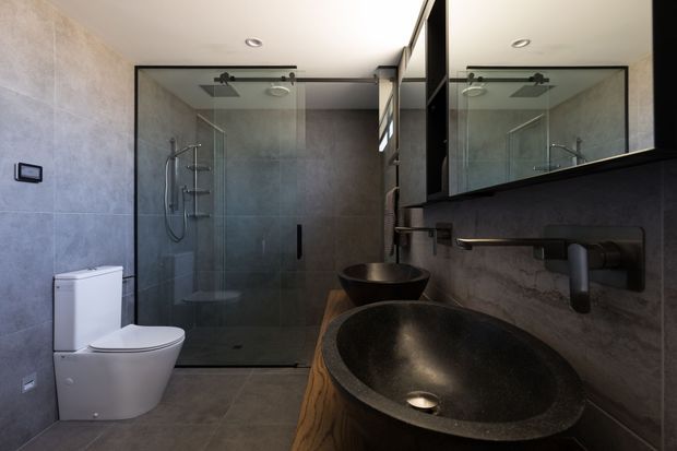 Contemporary bathroom with large-format grey stone-look wall and floor tiles, a frameless glass walk-in shower with black hardware, and two dark vessel basins set on a warm timber vanity. A large mirrored cabinet and matte black tapware create a refined, moody finish.