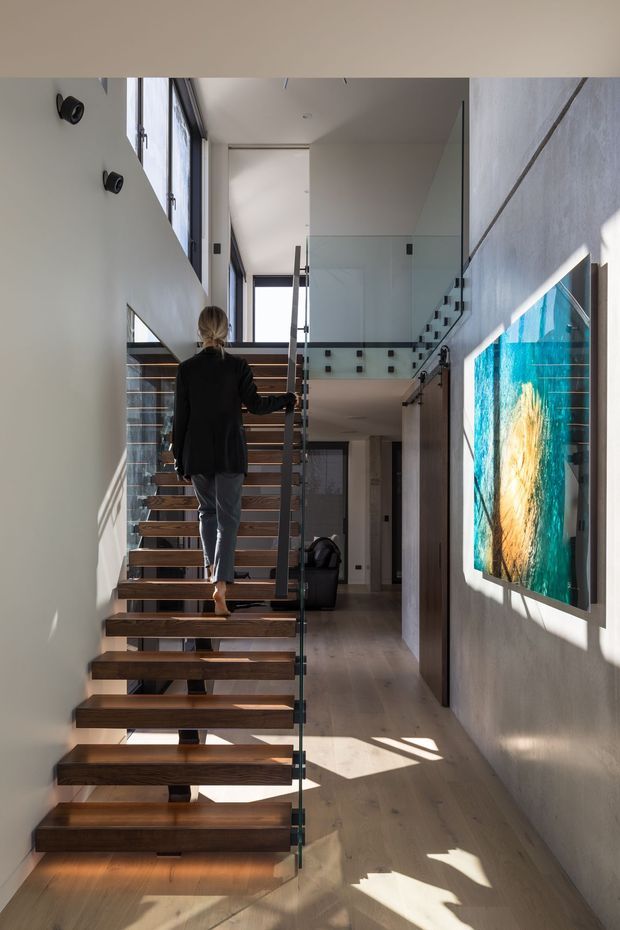 A bright, contemporary interior stairway with floating timber treads, slim dark metal supports, and clear glass balustrades. The stair rises through a tall, light-filled hallway with large vertical windows, pale walls, light timber flooring, and a colourful abstract artwork on the right wall.