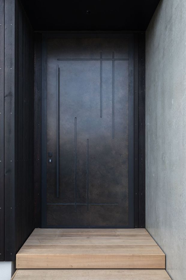 A close-up view of a dark, minimalist exterior entryway featuring a tall charcoal metal-clad front door with an abstract linear relief pattern, set within dark vertical timber or metal framing. The entry is approached by a small flight of pale timber steps, with a textured light-grey rendered wall on the right and a dark ceiling overhang above.