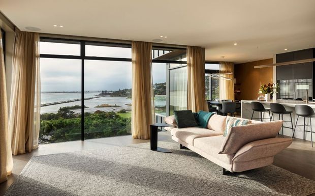 A bright, open-plan contemporary living room with a large cream sectional sofa, textured light rug, black side table, and full-height black-framed glazing overlooking a coastal waterfront. The living area flows into a modern kitchen with a dark island, bar seating, timber-toned cabinetry, and pendant lighting. Floor-to-ceiling curtains soften the expansive windows.