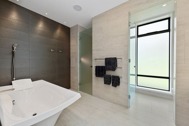 Contemporary bathroom with a freestanding white bathtub, frameless glass shower enclosure, dark textured feature wall, pale stone-look wall and floor tiles, heated towel rail with dark towels, and a large frosted window. Recessed ceiling lighting creates a clean, refined atmosphere.