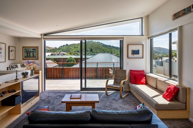 Bright contemporary living room opening onto a timber-decked balcony through extensive sliding glass doors. The room features a built-in window seat sofa with red cushions, black leather seating in the foreground, a timber coffee table, a rocking chair, low timber cabinetry, artwork, and broad views toward a green hillside and neighbouring rooftops. A high horizontal clerestory window and clean white surfaces enhance the airy, open feel.