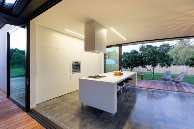 Bright contemporary open-plan kitchen with extensive white built-in cabinetry, a central island with cooktop and seating, polished concrete flooring, and large black-framed sliding glass doors opening to a timber deck and landscaped outdoor area. Linear recessed lighting and a sculptural rangehood contribute to the clean modern design.