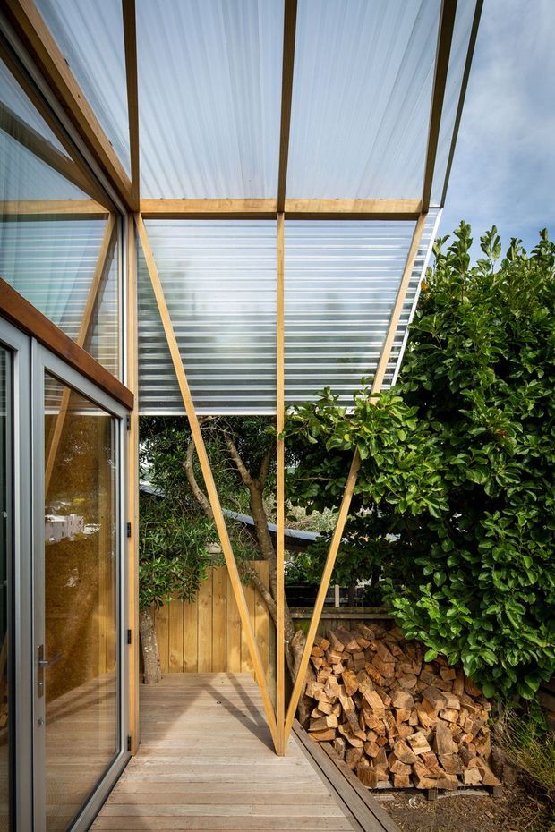 A narrow covered outdoor passage beside a building, featuring a translucent corrugated roof supported by exposed timber framing and angled braces. Timber decking continues along glazed timber-framed doors and walls, opening onto a leafy garden with a timber fence and stacked firewood. The image has a warm, natural, utilitarian architectural character with strong vertical and diagonal framing.