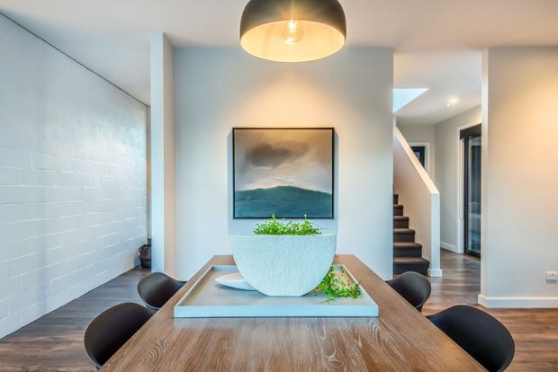 A contemporary dining area with a large timber dining table, dark rounded chairs, a central planter, and a framed landscape artwork. The room features white painted brickwork, smooth white walls, dark timber flooring, a sculptural pendant light, and an open hallway with a staircase in the background.