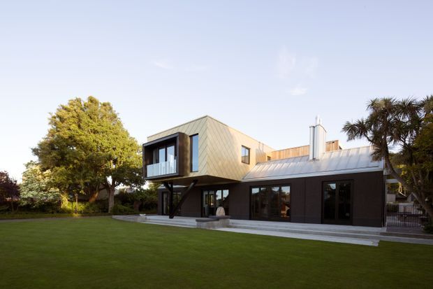 Exterior view of a contemporary two-storey residential house set beside a broad manicured lawn. The building combines a dark lower level with a projecting upper volume clad in light metal panels, extensive dark-framed glazing, and a low-pitched standing-seam metal roof. Mature trees and landscaped grounds surround the home.