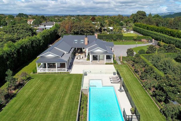 Aerial view of a large single-storey suburban residence with a dark grey pitched roof, covered outdoor verandah, expansive manicured lawns, landscaped gardens, paved entertaining terrace and a long rectangular swimming pool. The property is surrounded by mature hedges and trees, with distant hills visible beyond.