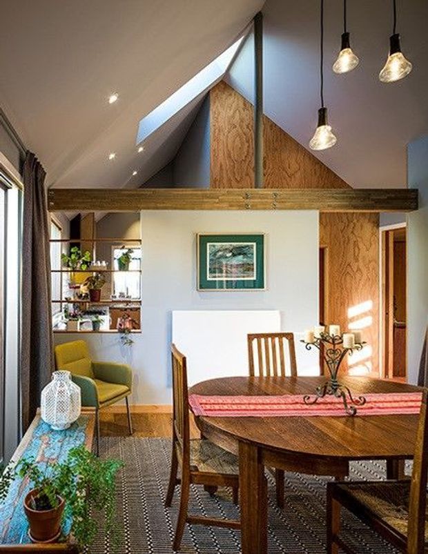 A warm, intimate dining room beneath a steeply pitched vaulted ceiling. A large round dark-wood dining table with wooden chairs dominates the foreground, with a pink table runner and candleholder. Exposed timber beams cross the room, while a timber-clad triangular gable and skylight bring strong architectural character. The room also includes a small green armchair, patterned rug, open shelving with plants, framed artwork, and pendant lighting.