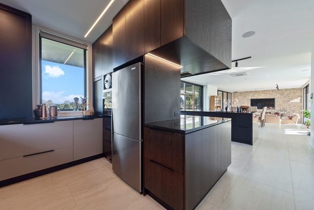 A contemporary open-plan kitchen with dark timber cabinetry, a large central island, integrated stainless-steel refrigerator, pale tiled flooring, and linear LED lighting. The kitchen opens toward a bright dining and living area, with extensive glazing and a stone-textured feature wall visible in the background.