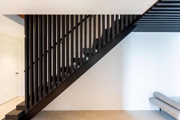 A minimalist interior stairway with a black timber or metal balustrade and dark stair structure set against crisp white walls. The staircase runs diagonally across the room, with a light neutral floor and part of a pale sofa visible at right. A slatted ceiling or stair underside detail adds strong linear contrast.