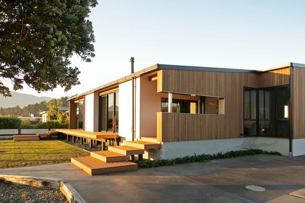 A single-storey contemporary house viewed from the front, with a low flat roof, warm timber-clad feature walls, white rendered wall sections, large dark-framed windows and doors, and a raised timber deck with broad steps. The home sits on a landscaped suburban site with a paved driveway and lawn.