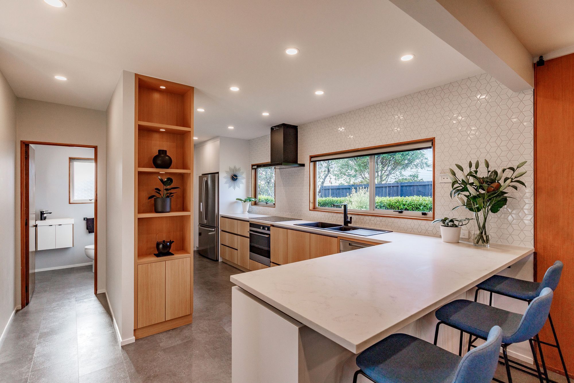 The shelving unit conceals built-in cabinets. The blend of wood, white, and black accents creates a timeless and inviting ambiance, while the textured splashback tiles add subtle interest.