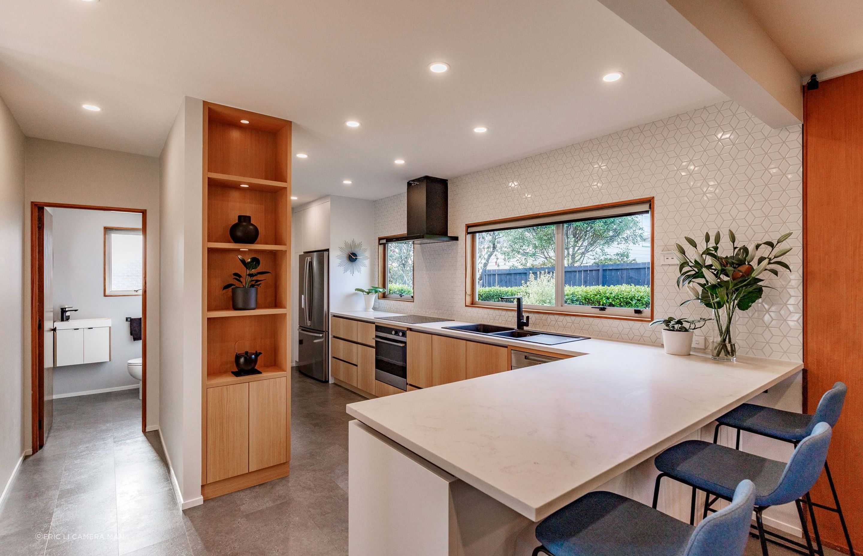 The shelving unit conceals built-in cabinets. The blend of wood, white, and black accents creates a timeless and inviting ambiance, while the textured splashback tiles add subtle interest.