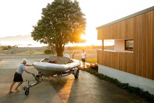 Exterior view of a contemporary single-storey building with a long low-pitched roof and warm vertical timber cladding over a white rendered base. A paved driveway or forecourt occupies the foreground, with a boat trailer being moved beside the building. Large glazing and an open covered edge connect the building to the outdoor area. The setting appears coastal or waterfront, with bright low sunlight and distant water or shoreline visible beyond the site.