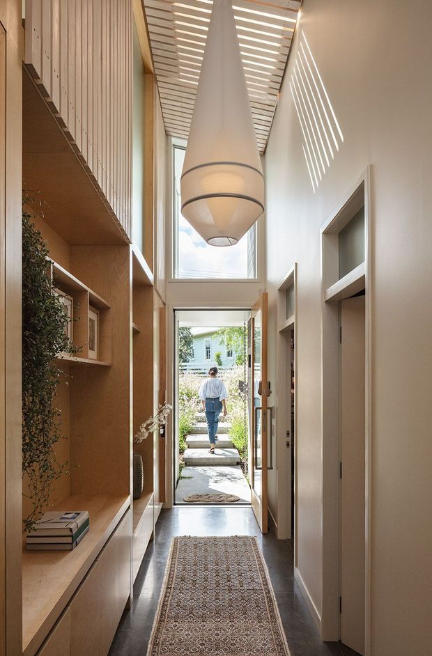 A narrow, tall residential entry hall with a long patterned runner, dark polished flooring, pale walls, and extensive warm timber built-in shelving and cabinetry along the left side. A large sculptural pendant hangs beneath a slatted timber ceiling, casting linear sunlight shadows across the wall. Full-height glazing and an open door connect the hall to a planted outdoor path, while recessed doorways and joinery line the right wall.