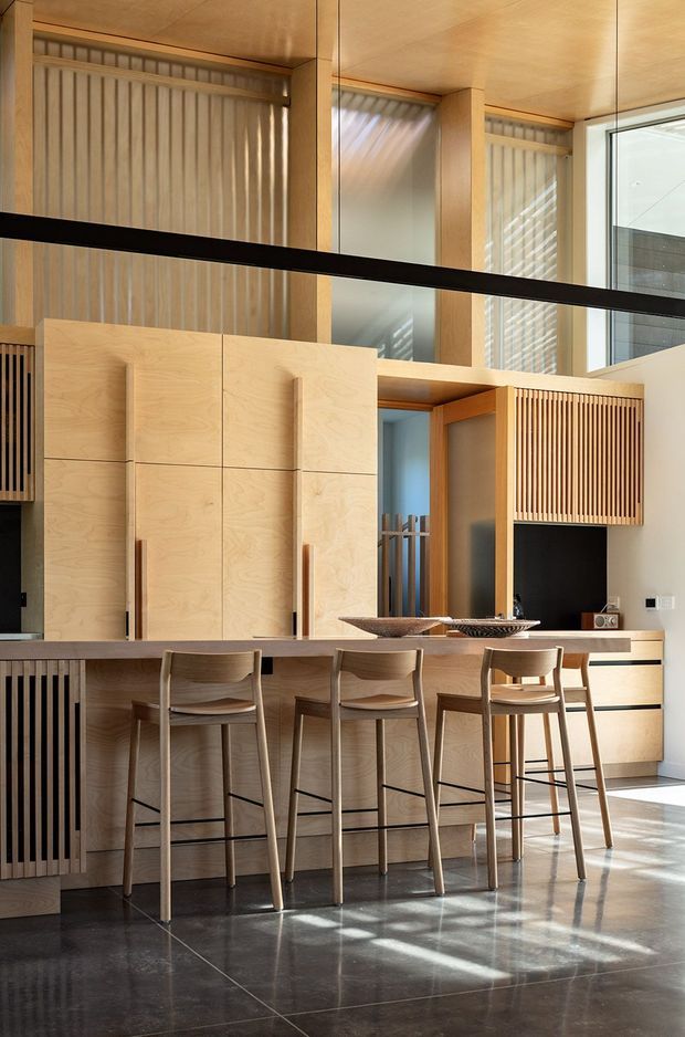 A warm, double-height contemporary kitchen with extensive light timber cabinetry and vertical slatted feature panels. A long island with a dark stone-looking benchtop provides seating for four on timber bar stools. Full-height glazing and clerestory-like upper windows bring in strong natural light, which reflects across the dark polished floor. The room includes integrated appliances and a black-framed internal or external glazed opening.