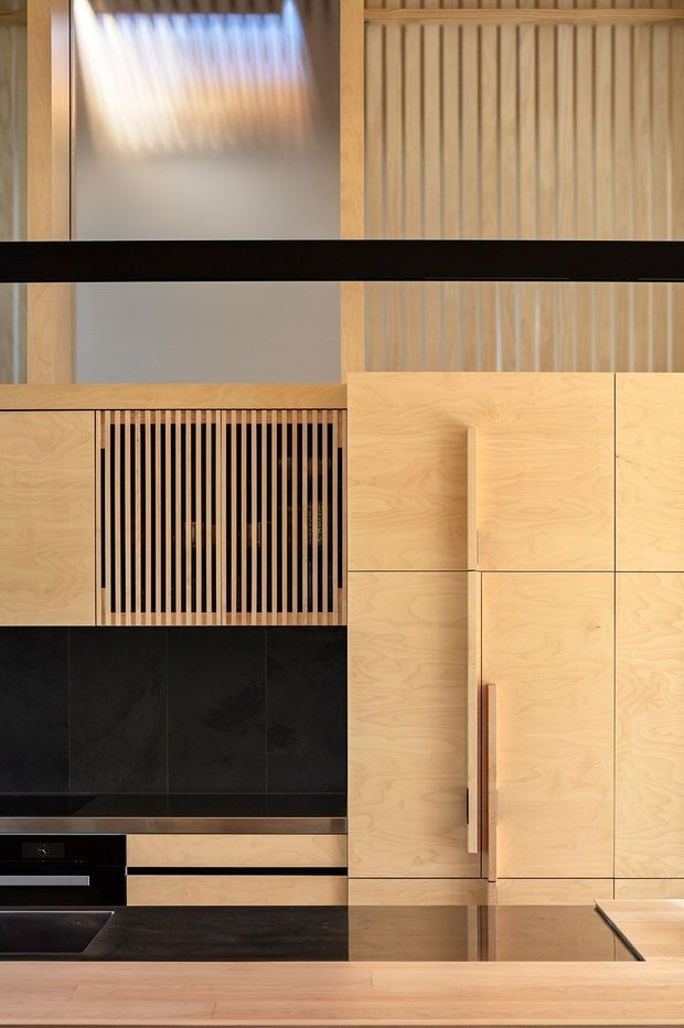 Close-up view of a refined contemporary kitchen featuring light timber cabinetry and integrated wood-grain handles, a dark stone-look splashback and benchtop, black integrated appliances, and a vertical timber slatted feature panel. A dark horizontal structural or decorative beam crosses the upper portion, with warm timber wall panelling and a softly lit reflective panel behind.
