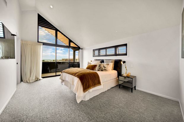 Bright contemporary bedroom with a large upholstered bed, layered neutral bedding, dark bedside tables, grey carpet, white walls, and expansive black-framed glazing. High-level louvred windows and an angled glazed section bring in abundant daylight, with curtains opening toward an exterior view.