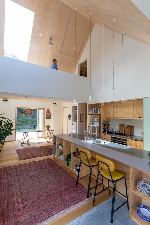 Bright open-plan kitchen and living space with a large island and raised breakfast bar, timber cabinetry, stainless-steel benchtops, and three bar stools. The room has a dramatic double-height ceiling lined with warm plywood, exposed pendant lights, a mezzanine or upper-level opening, timber floors, patterned rugs, and large glazing opening toward greenery.