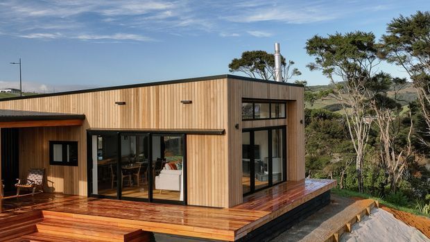 Exterior view of a compact contemporary timber-clad house set among coastal vegetation. The single-storey building has a flat roof, extensive dark-framed glazing, a broad timber deck, and a raised structure overlooking the surrounding landscape.