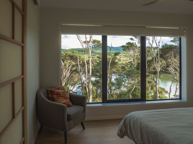 A calm bedroom interior with a large floor-to-ceiling picture window overlooking a wooded, leafy landscape and water beyond. The room features a grey upholstered armchair with a patterned cushion, a partially visible bed with a neutral textured cover, pale walls, light timber flooring, and dark-framed glazing. A simple linear wall detail or built-in paneling is visible along the left side.