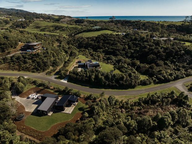 Aerial view of a sparsely developed, heavily vegetated rural or semi-rural coastal landscape, with several contemporary detached homes on large landscaped lots, winding paved roads, rolling pasture, dense native vegetation, and the ocean visible across the horizon.