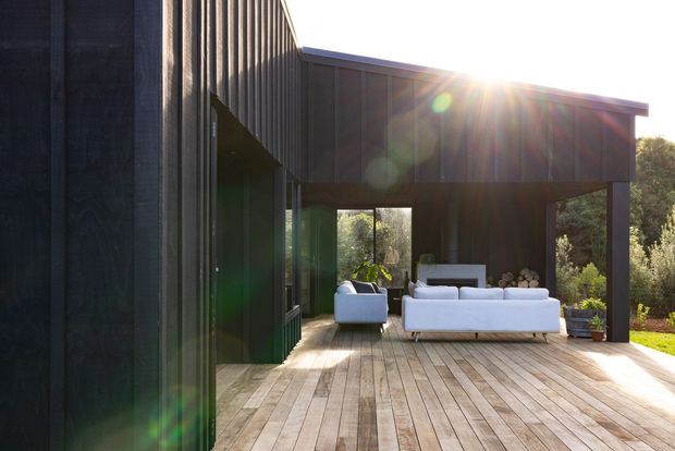 Exterior view of a contemporary dark-clad house opening onto a spacious timber deck. The covered outdoor area contains white lounge seating and a small outdoor fireplace or hearth, with extensive glazing and surrounding greenery visible beyond. Strong sunlight creates lens flare and highlights the indoor-outdoor connection.