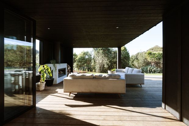 A contemporary indoor-outdoor living room opening onto a timber deck and landscaped garden. The room has a dark timber-lined ceiling and walls, extensive floor-to-ceiling glazing and large glazed doors, timber flooring/decking, a pale sofa and armchair, and a built-in fireplace surround. Strong sunlight creates long shadows across the floor, giving the space a warm, relaxed connection to the outdoors.