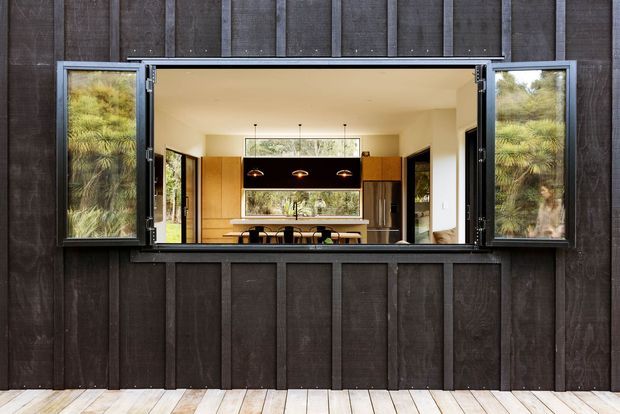 Exterior view of a dark timber-clad building with a large open serving-style window. Through the opening, a warm contemporary kitchen or dining space is visible, featuring light timber cabinetry, a dark overhead island or joinery element, pendant lights, stools, and extensive views toward greenery. A pale timber deck runs along the base of the facade.