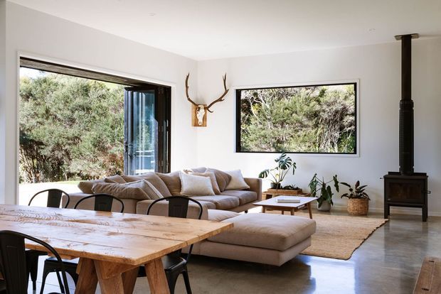 Bright open-plan living and dining area with large windows overlooking dense greenery. The room features a low beige sectional sofa and ottoman, a rustic solid timber dining table with dark chairs, polished concrete flooring, white walls and ceiling, indoor plants, and a black freestanding wood-burning stove with flue. A mounted antler decoration adds a rustic natural accent.