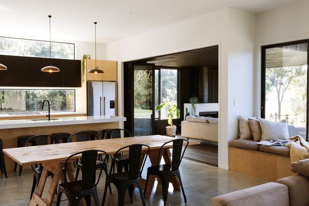 Open-plan contemporary kitchen, dining and living area with a long timber dining table, black metal dining chairs, a white island bench, dark kitchen cabinetry, pendant lights and built-in window seating. Large glazed openings connect the bright interior to a leafy outdoor setting, while a dark-framed opening leads into the living room.