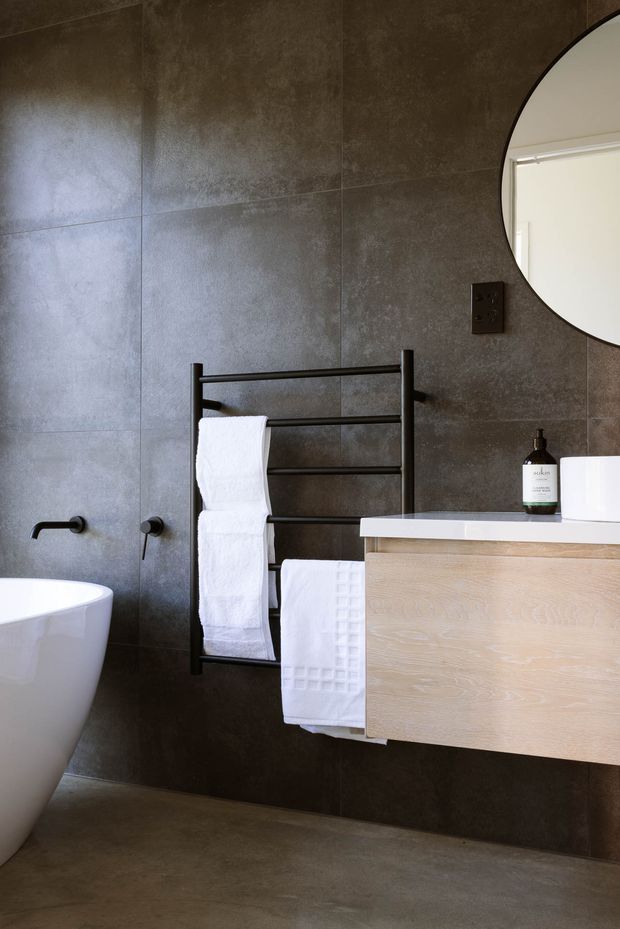 A contemporary bathroom with large dark grey textured wall tiles, a grey floor, a white freestanding bathtub, wall-mounted black tapware, a black heated towel rail with white towels, and a floating light timber vanity with a white benchtop. A large round mirror and dark electrical fittings complete the space.