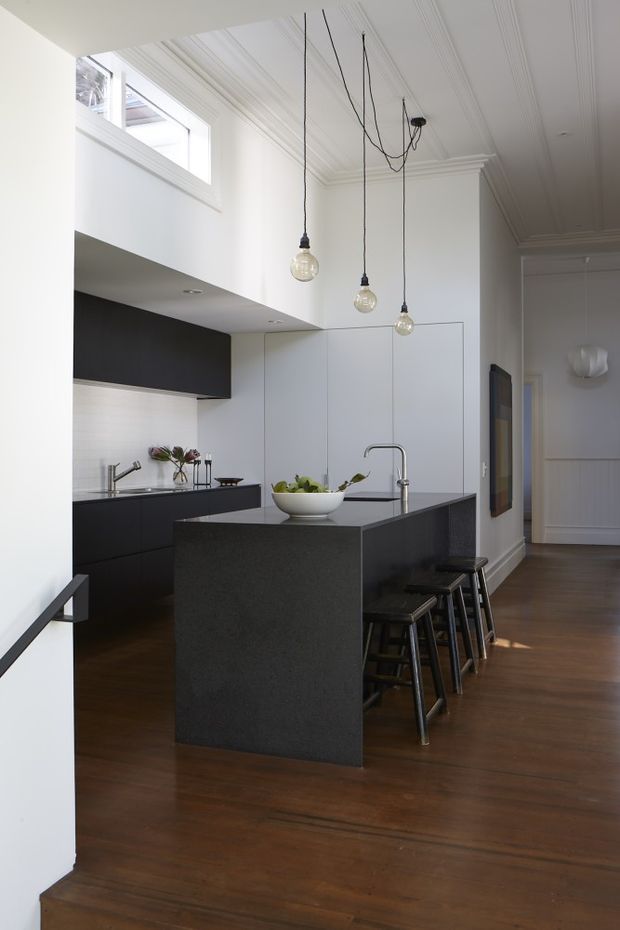 A bright contemporary kitchen viewed from an adjoining hallway or living area. The room features a large dark stone island with integrated seating, matching dark cabinetry, white walls and cabinetry, timber flooring, a stainless-steel tap, and three suspended globe pendant lights. High-level glazing and detailed white ceiling mouldings add architectural character, while the adjacent hallway continues to the right.
