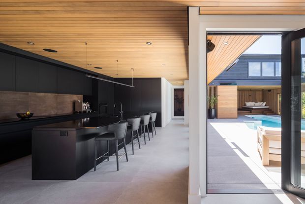 Contemporary open-plan kitchen with a long black island and integrated cabinetry, dark stone-look splashback, bar seating, and a warm timber slatted ceiling. Large glazed doors open directly to a bright courtyard and swimming pool area, with additional timber-clad exterior elements visible beyond.
