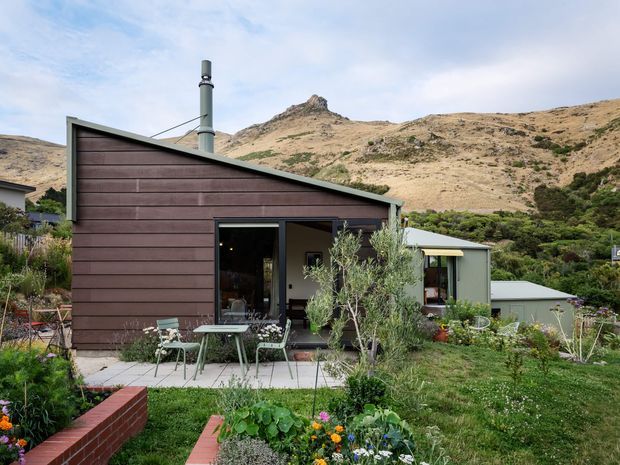 Exterior view of a compact single-storey house in a mountainous setting. The home has dark horizontal timber cladding, a distinctive sloping roofline, metal chimney, glazed doors, and a small paved outdoor sitting area surrounded by landscaped garden beds. Rugged dry hills and native vegetation rise behind the property.